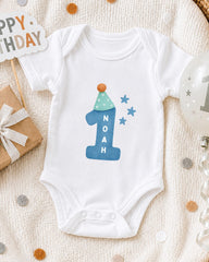 A white onesie with a blue number one and stars on it, along with a "Happy Birthday" sign and a birthday balloon, all placed on a white blanket with confetti.