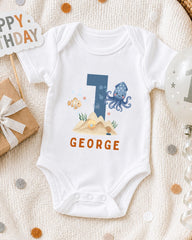 A white onesie with a blue octopus and the name "GEORGE" printed on it, along with a "1" and a "Happy Birthday" sign.
