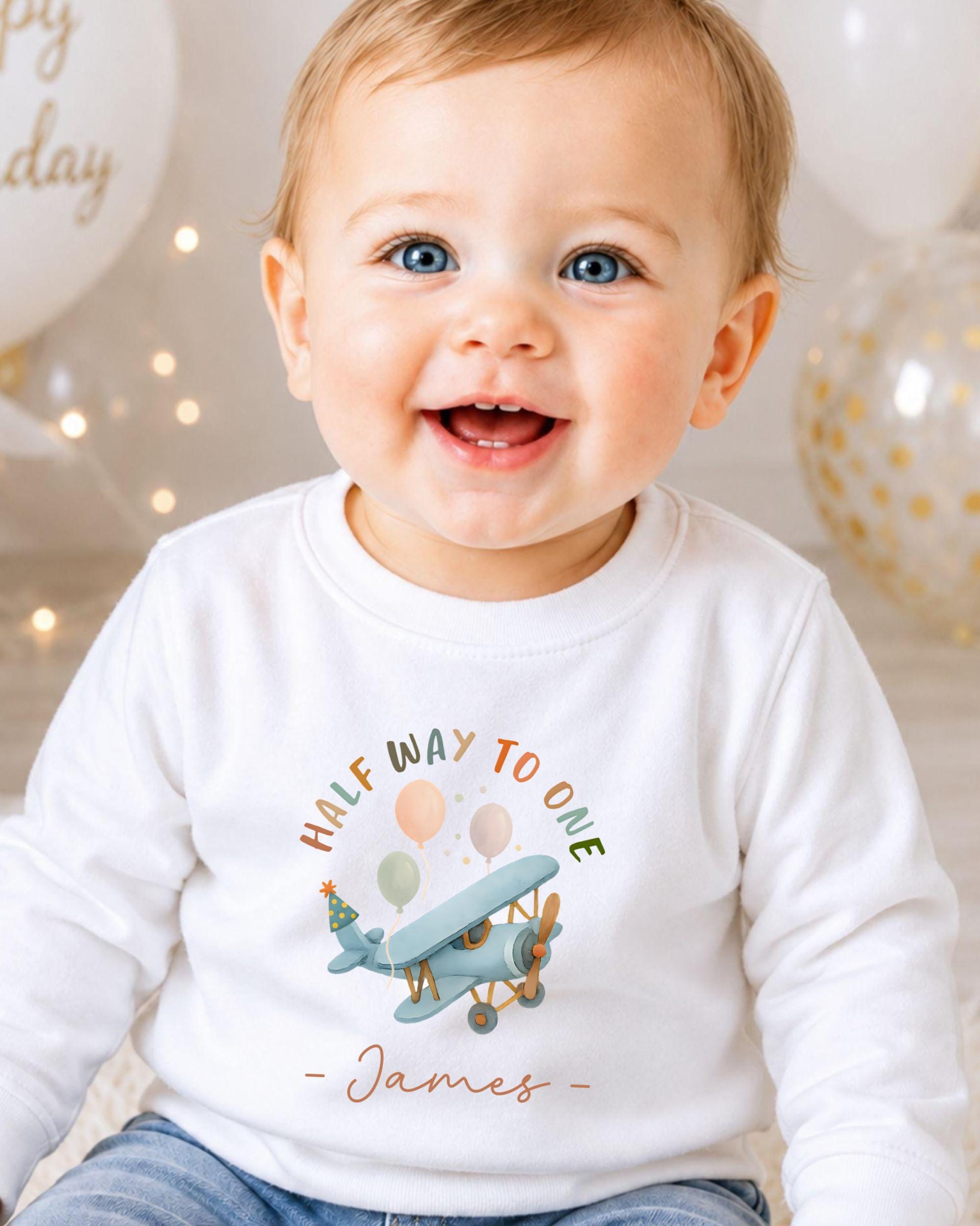 A smiling baby boy wearing a white t-shirt with a graphic of a plane and the text "Half way to one" and "James" printed on it.