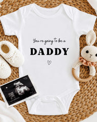 A white baby onesie with the text "You're going to be a DADDY" printed on it, along with a cute stuffed bunny toy and a black and white photograph of an Ultrasound image.