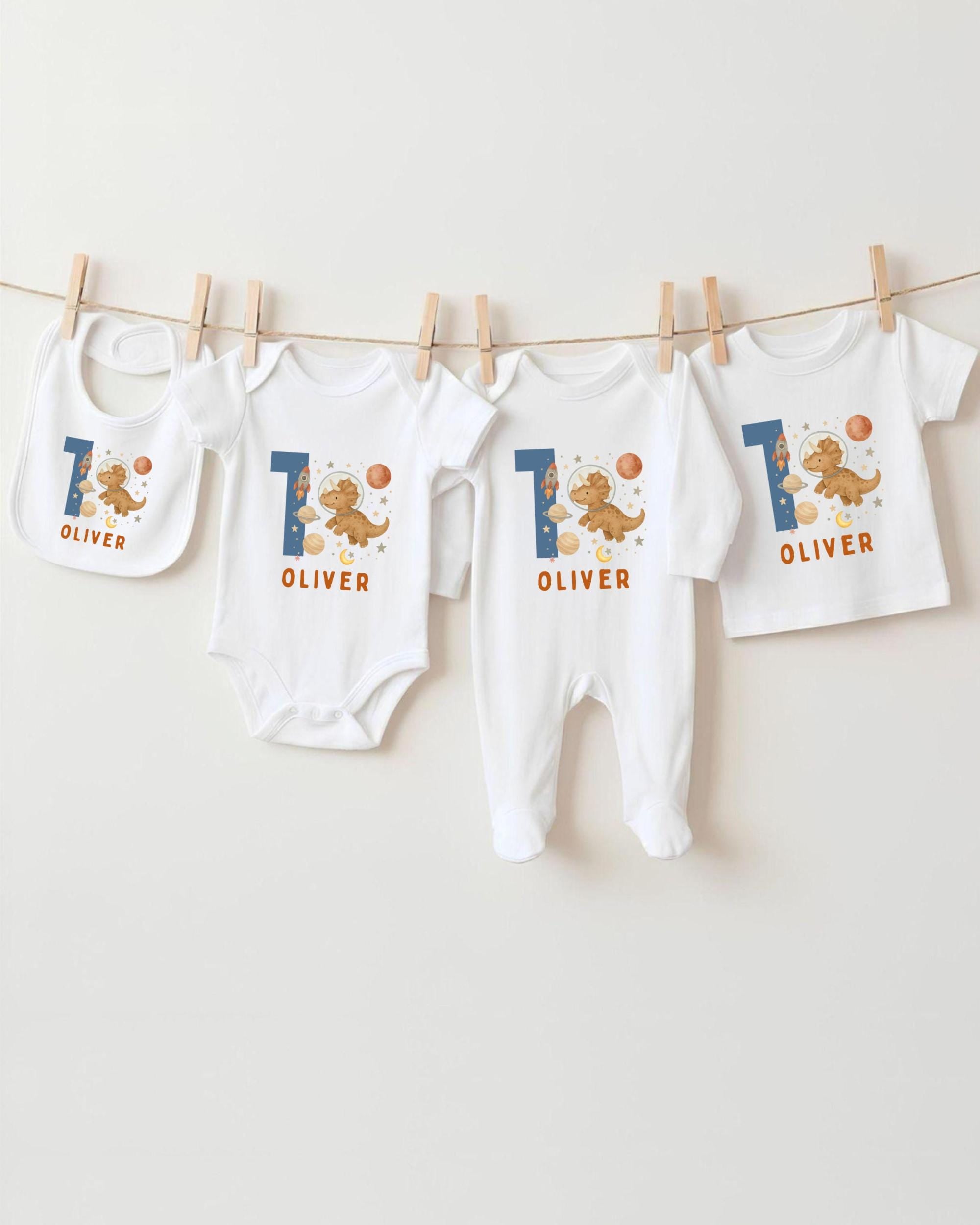 A collection of white baby clothes, including onesies and bibs, with the name "OLIVER" printed on them. The clothes are hanging on a clothesline, and there are four of them in total.