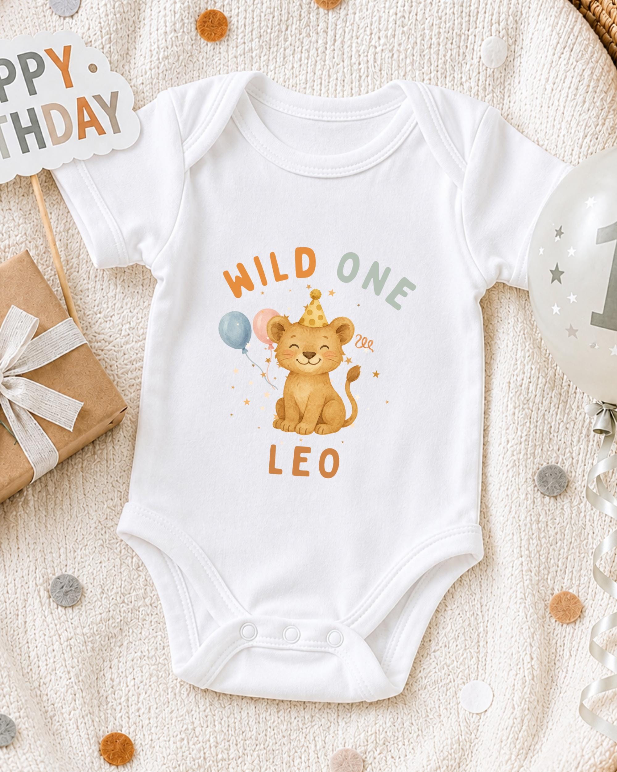 A white onesie with a cartoon lion and the text "Wild One Leo" printed on it, along with a birthday sign and balloons, suggesting a celebration for a baby's first birthday.