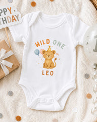 A white onesie with a cartoon lion and the text "Wild One Leo" printed on it, along with a birthday sign and balloons, suggesting a celebration for a baby's first birthday.