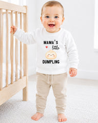 A smiling baby boy wearing a white sweatshirt with a cartoon image of a dumpling and the text "Mama's little DUMPLING" standing in front of a wooden crib.