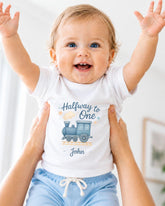A smiling baby wearing a white t-shirt with a train graphic and the text "Halfway to One John" is being held up by someone's arms.