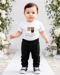 A young child wearing a white t-shirt with a graphic of two bears and text, standing on a white carpet in front of a floral backdrop.