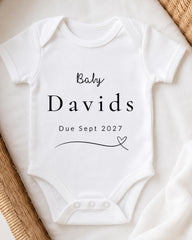 A white baby onesie with the text "Baby Davids Due Sept 2027" printed on it.