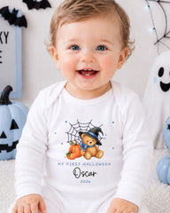 A smiling baby boy wearing a white onesie with a Halloween-themed design, including a teddy bear and pumpkins, is sitting in front of Halloween decorations.