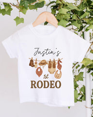 A white t-shirt with a graphic design featuring a cowboy hat, boots, and other western-style clothing items, along with the text "Justin's 1st Rodeo".