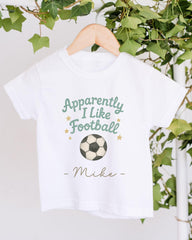 A white t-shirt with a soccer ball graphic and text that says "Approximately I Like Football - Mike" hanging on a wooden hanger.
