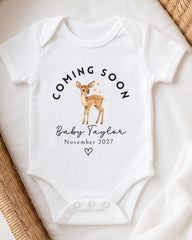 A white baby onesie with a graphic of a deer and the text "Coming soon Baby Taylor November 2027" printed on it.