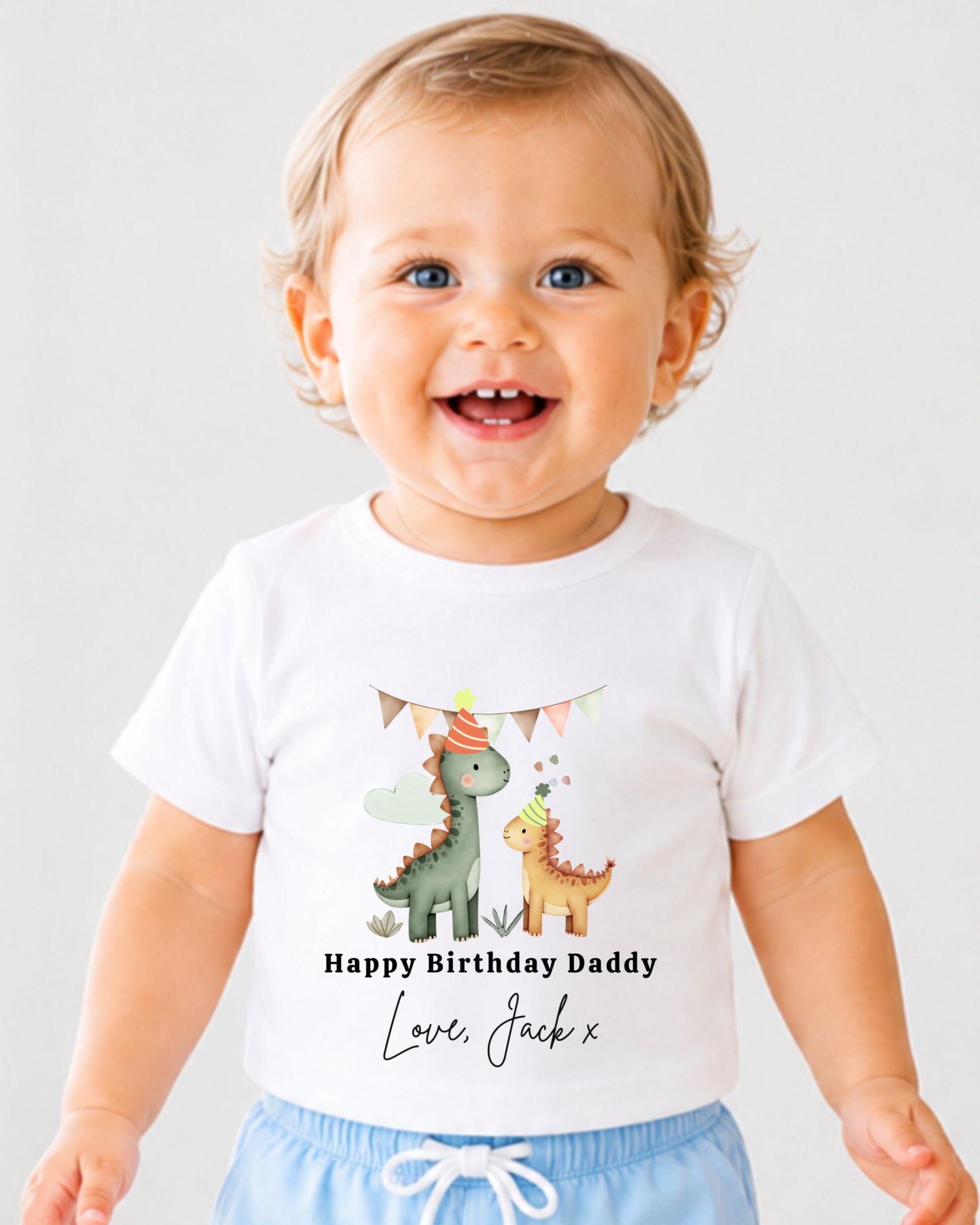 A smiling baby boy wearing a white t-shirt with a dinosaur graphic and text that says "Happy Birthday Daddy Love Jack X".