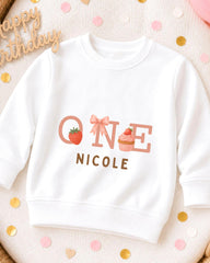 A white sweatshirt with a personalized text and graphics design, including a strawberry, cupcake, and the text "ONE NICOLE".