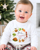 A smiling baby wearing a white shirt with a personalized Christmas-themed design, sitting in front of a Christmas tree.