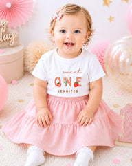 A smiling baby girl wearing a white t-shirt with the text "Sweet One" and a pink skirt, sitting on a pink and white background with balloons and confetti.
