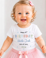 A smiling baby girl wearing a white t-shirt with text and a pink bow, standing in front of a white background.