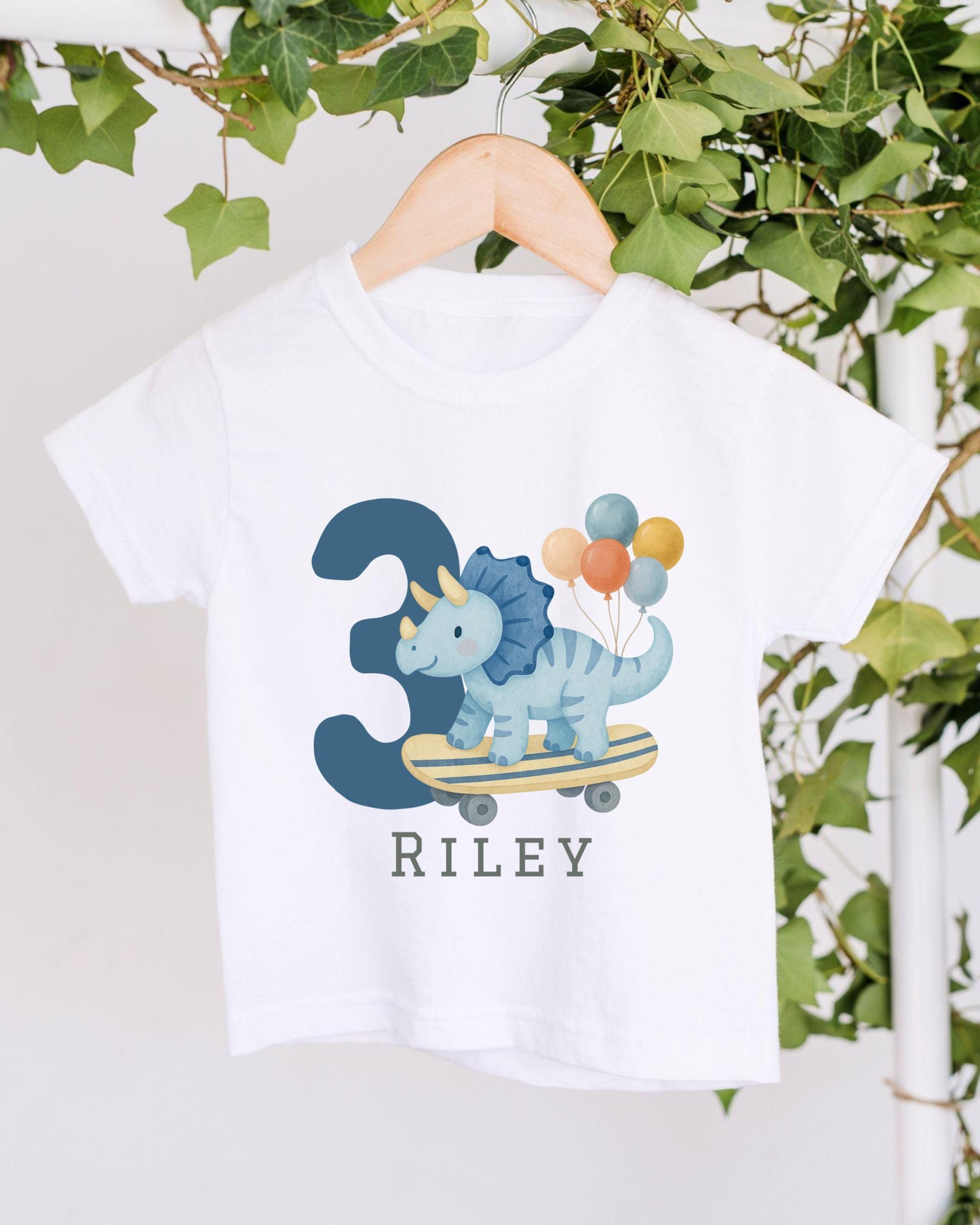 A white t-shirt with a graphic design of a blue dinosaur riding a skateboard, surrounded by colorful balloons, and the name "Riley" printed below.