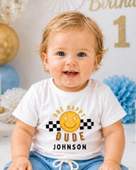 A smiling baby boy wearing a white t-shirt with a happy face and text, sitting in front of a birthday party backdrop.
