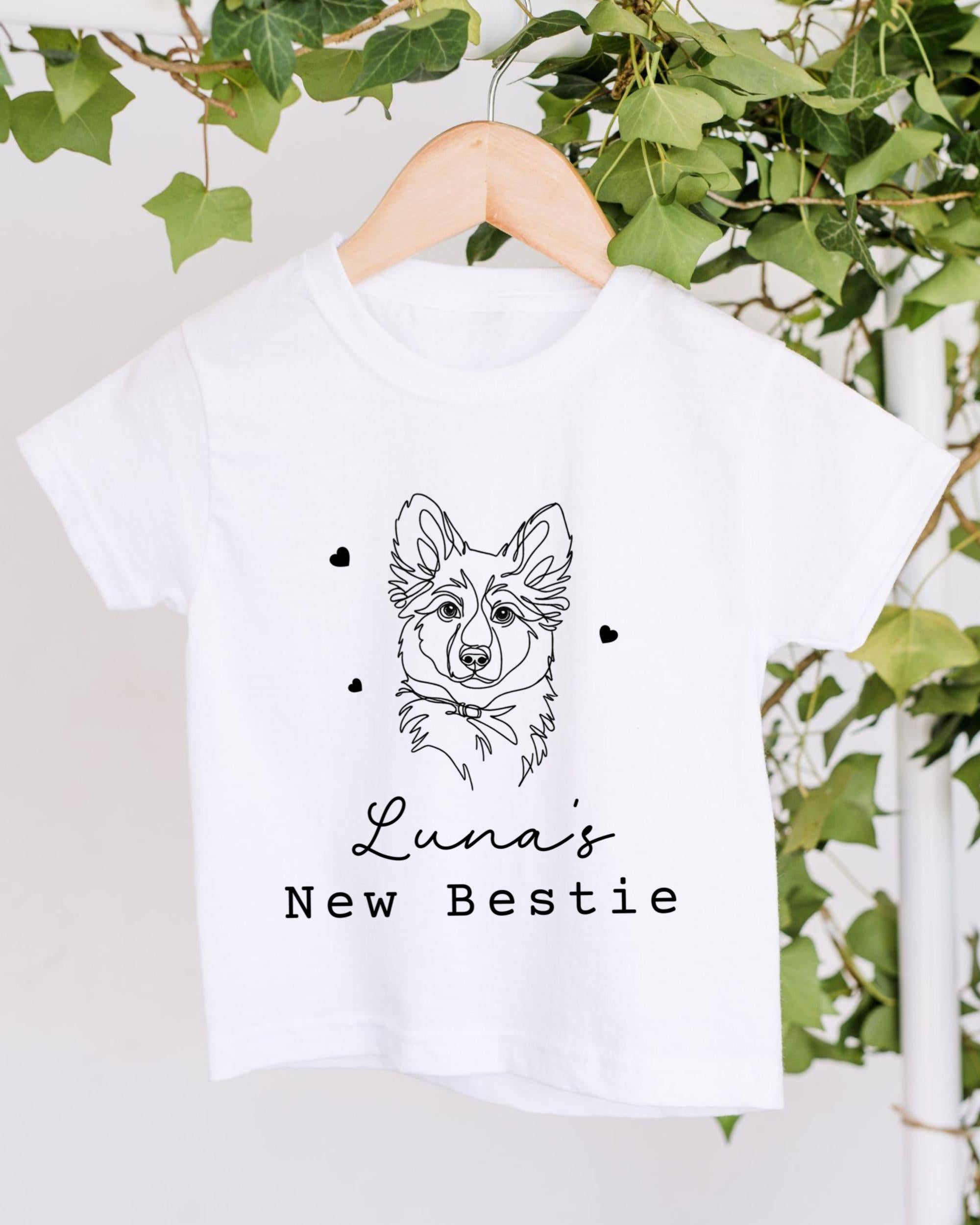 A white t-shirt with a black graphic of a dog's face and the text "Luna's New Bestie" printed on it, hanging on a wooden hanger.