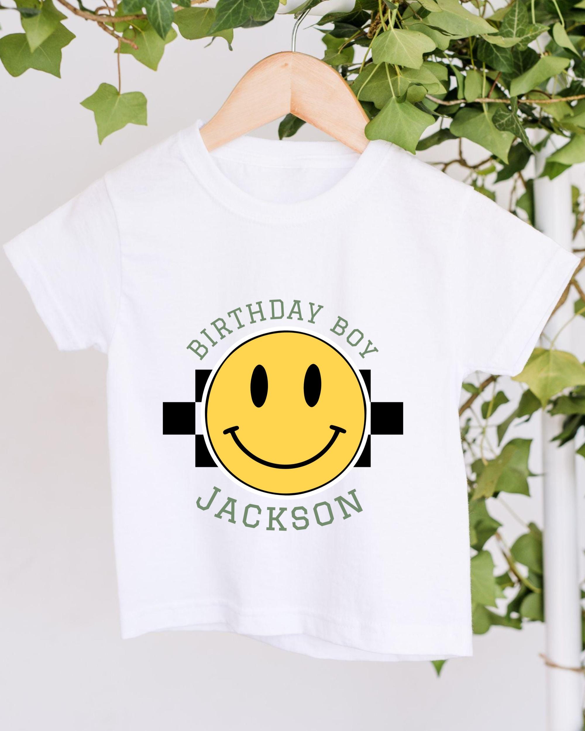 A white t-shirt with a yellow smiley face graphic and text printed on it, hanging on a wooden hanger.