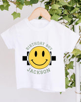 A white t-shirt with a yellow smiley face graphic and text printed on it, hanging on a wooden hanger.