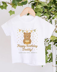 A white t-shirt with a graphic design featuring a teddy bear and the text "Happy Birthday Daddy!" printed on it, hanging on a wooden hanger.