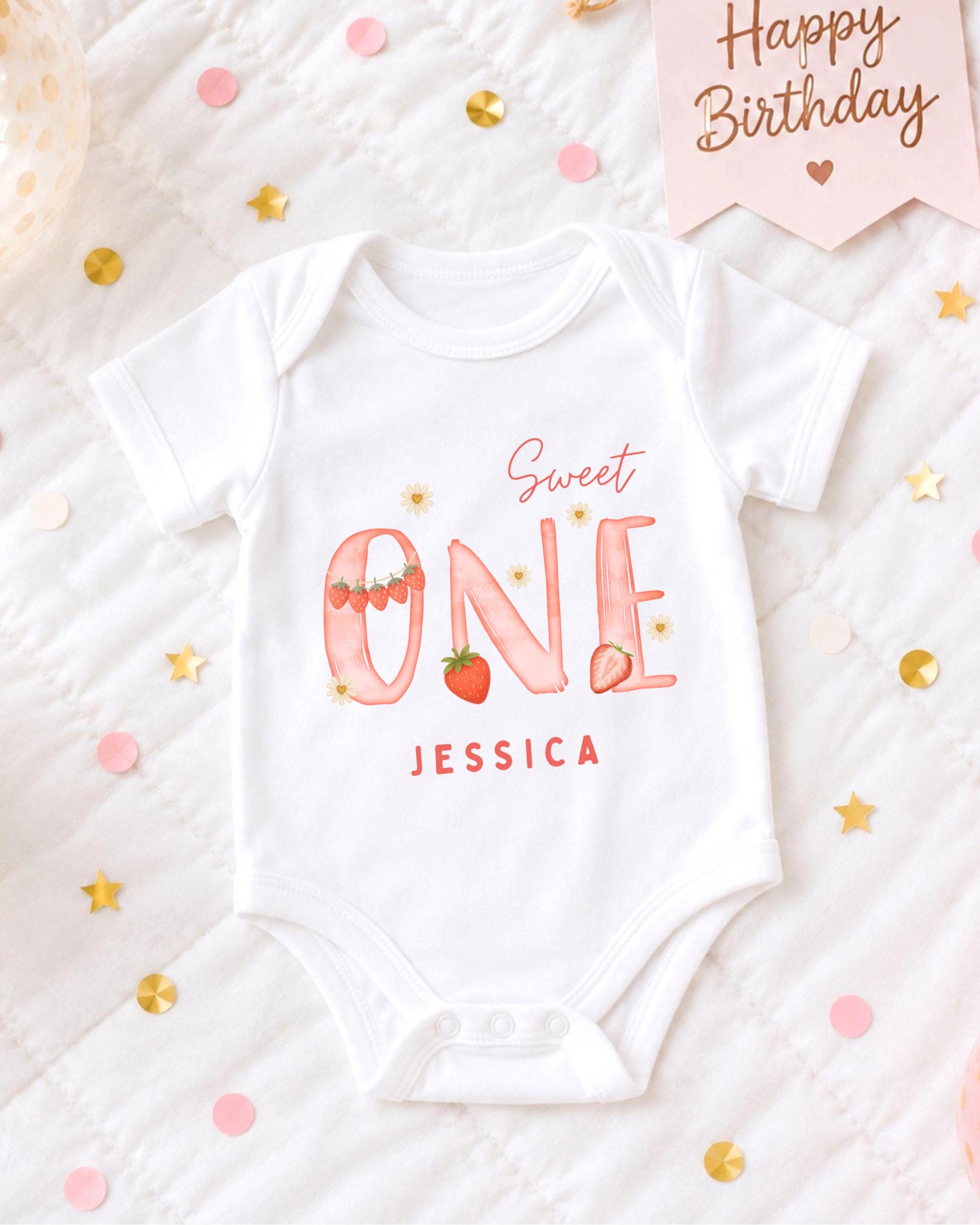 A white onesie with the text "Sweet One" and " Jessica" printed on it, along with a pink banner that says "Happy Birthday". The onesie is placed on a bed with confetti and stars scattered around it.