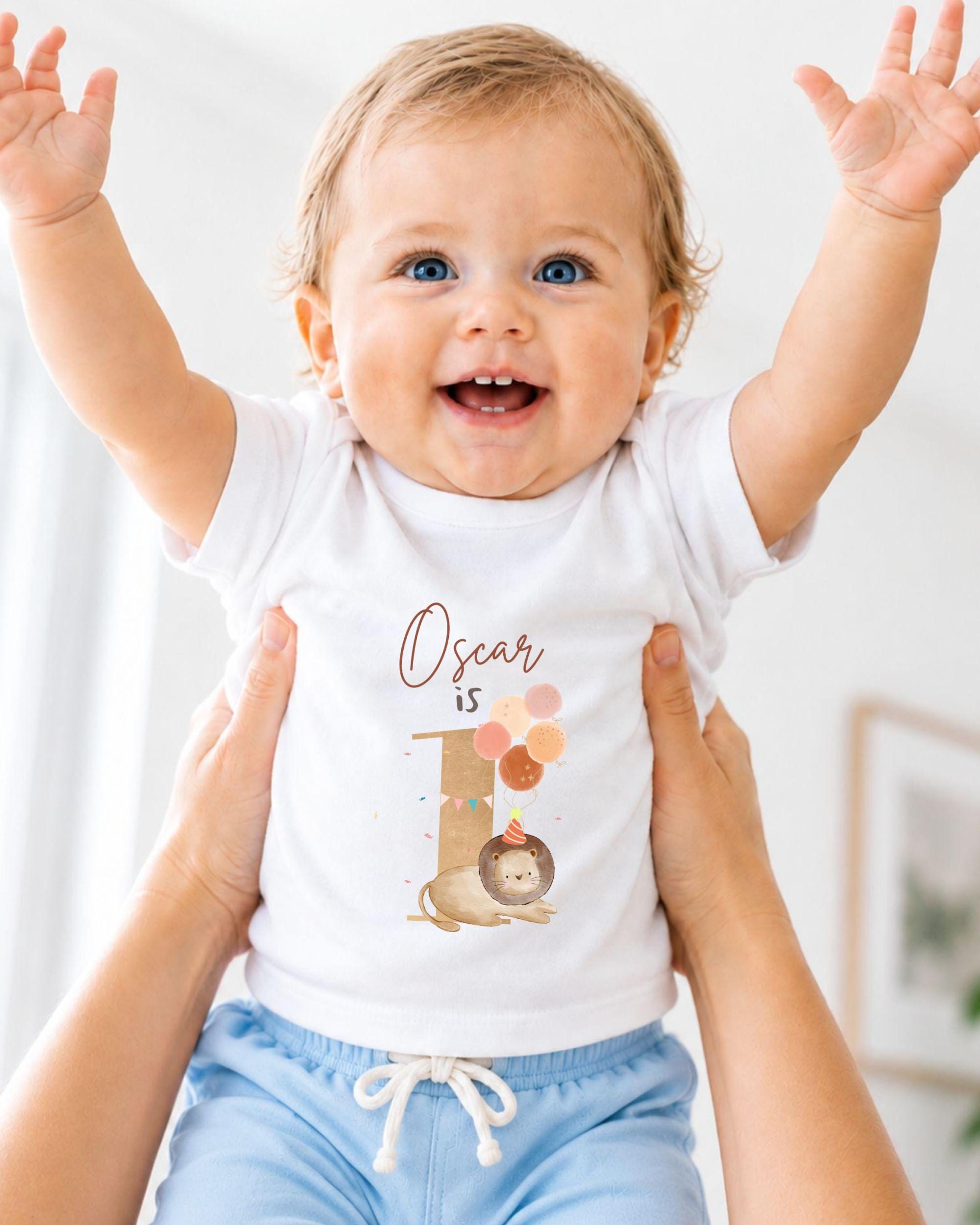 A smiling baby wearing a white t-shirt with a lion graphic and the text "Oscar is 1" is being held up by two people.