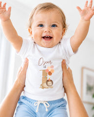 A smiling baby wearing a white t-shirt with a lion graphic and the text "Oscar is 1" is being held up by two people.