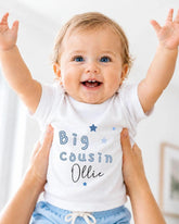 A smiling baby wearing a white shirt with the text "Big Cousin Ollie" is being held up by someone's arms.