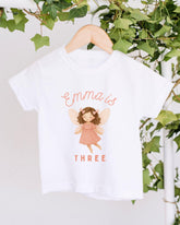 A white t-shirt with a graphic of a fairy and the text "Emma is THREE" printed on it, hanging on a wooden hanger.