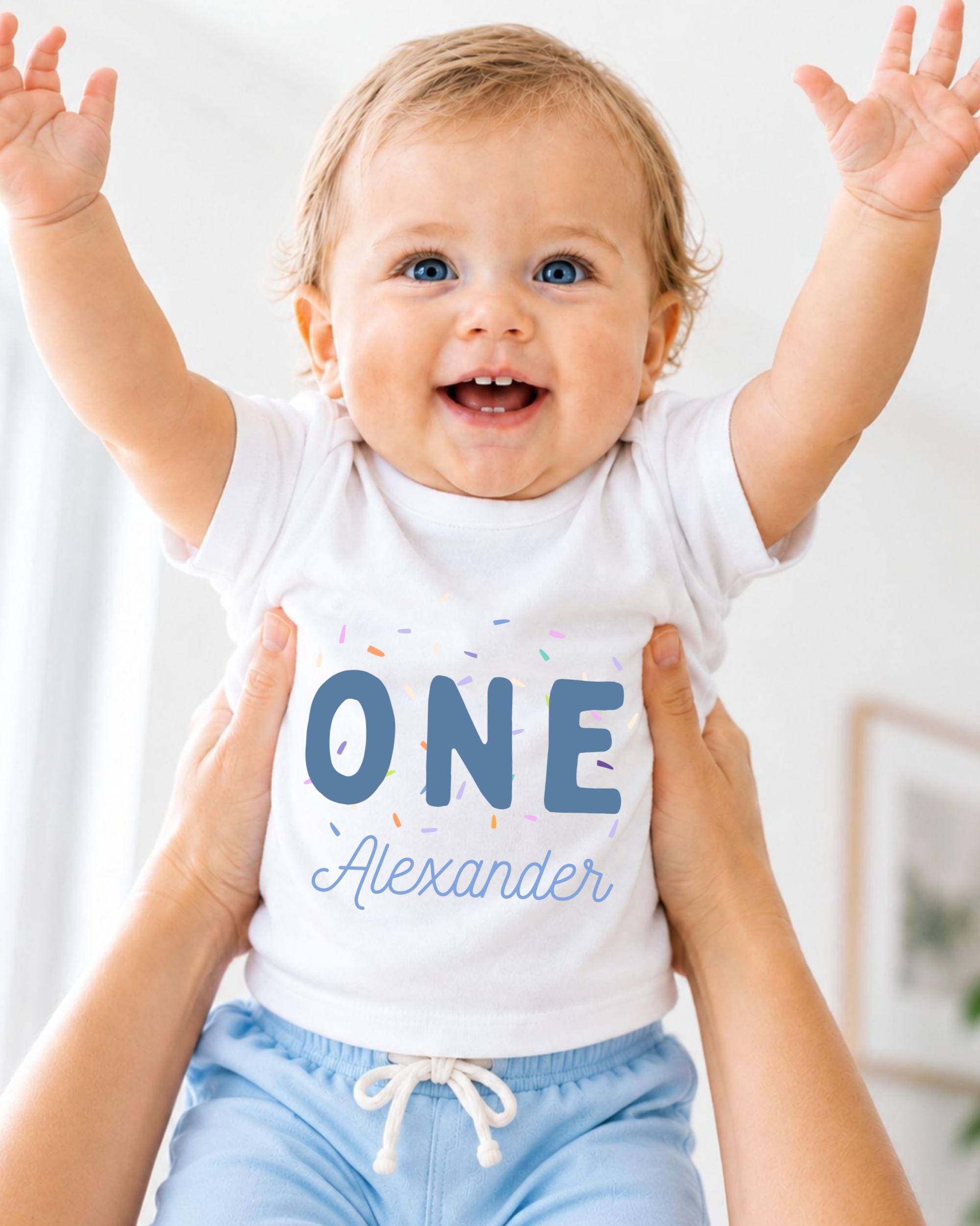 A smiling baby wearing a white shirt with the text "ONE Alexander" is being held up by someone's arms.