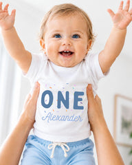 A smiling baby wearing a white shirt with the text "ONE Alexander" is being held up by someone's arms.