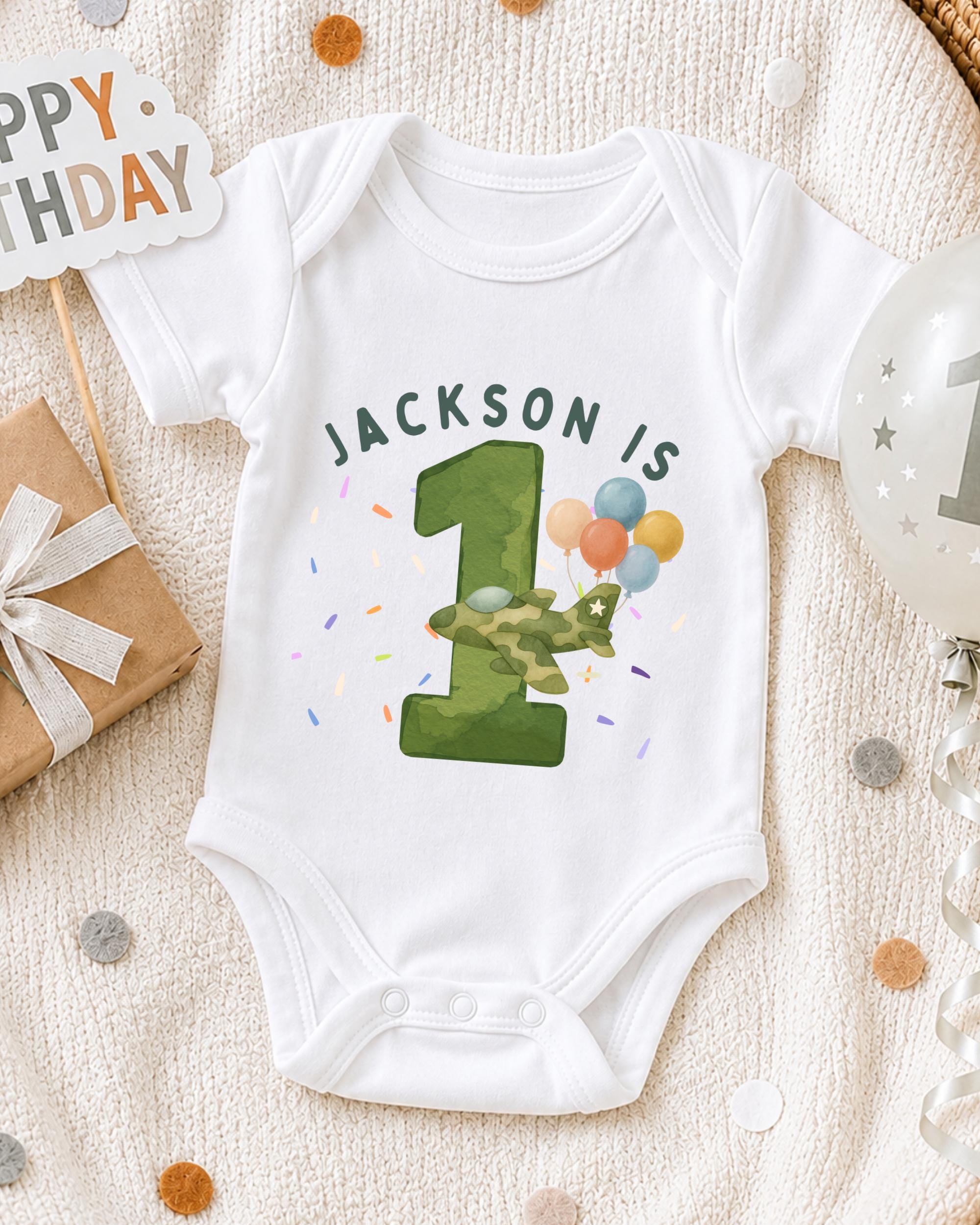 A white onesie with the text "JACKSON IS 1" printed on it, along with a green number one and colorful balloons, is displayed on a bed with a "Happy Birthday" sign and confetti.