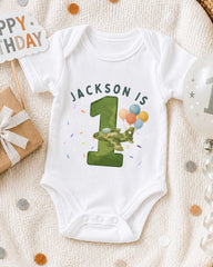 A white onesie with the text "JACKSON IS 1" printed on it, along with a green number one and colorful balloons, is displayed on a bed with a "Happy Birthday" sign and confetti.