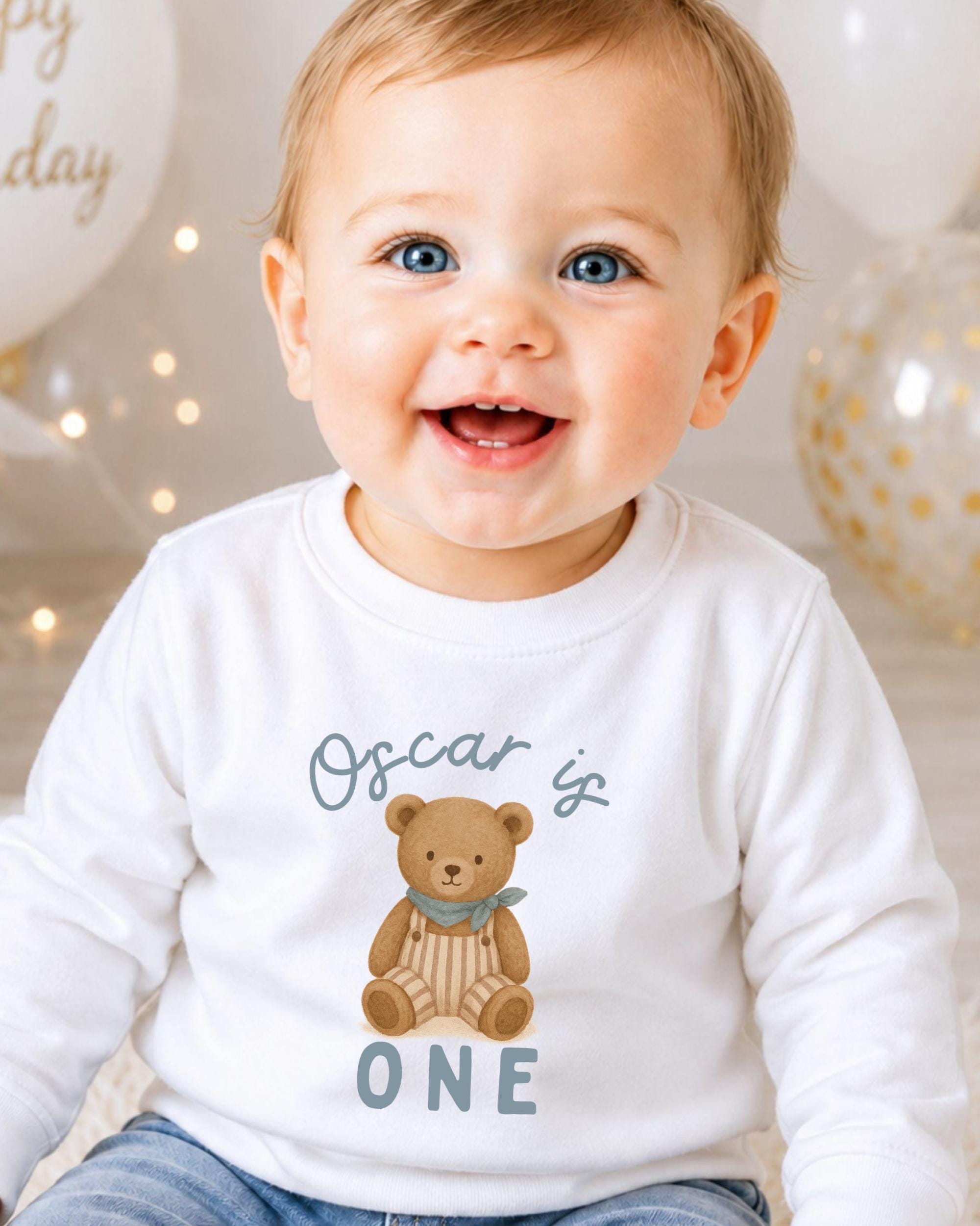 A smiling baby boy wearing a white shirt with a teddy bear graphic and the text "Oscar is 1" printed on it.