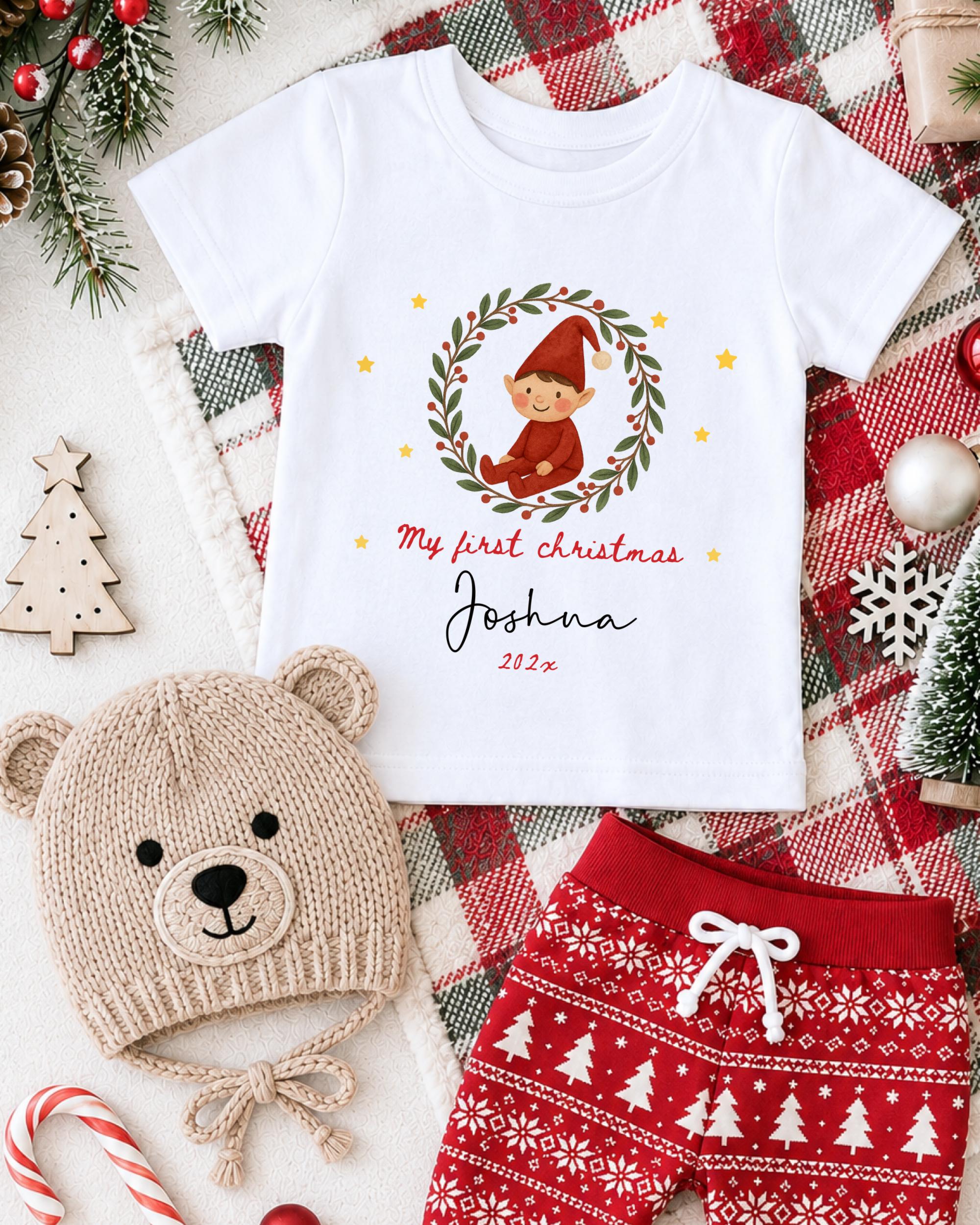 A white t-shirt with a Christmas-themed design, a beige knit hat with a Christmas tree, and red and white patterned shorts, all arranged on a plaid blanket with Christmas decorations.
