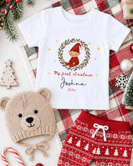 A white t-shirt with a Christmas-themed design, a beige knit hat with a Christmas tree, and red and white patterned shorts, all arranged on a plaid blanket with Christmas decorations.