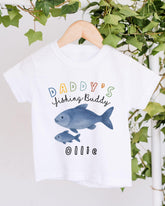 A white t-shirt with a colorful fish graphic and text that says "DADDY'S JIGGLING BUDDY OLLIE".
