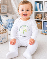 A smiling baby boy wearing a white onesie with a tennis racket and the name "Grayson" printed on it, sitting on a carpeted floor in a room with a bookshelf and stuffed animals in the background.
