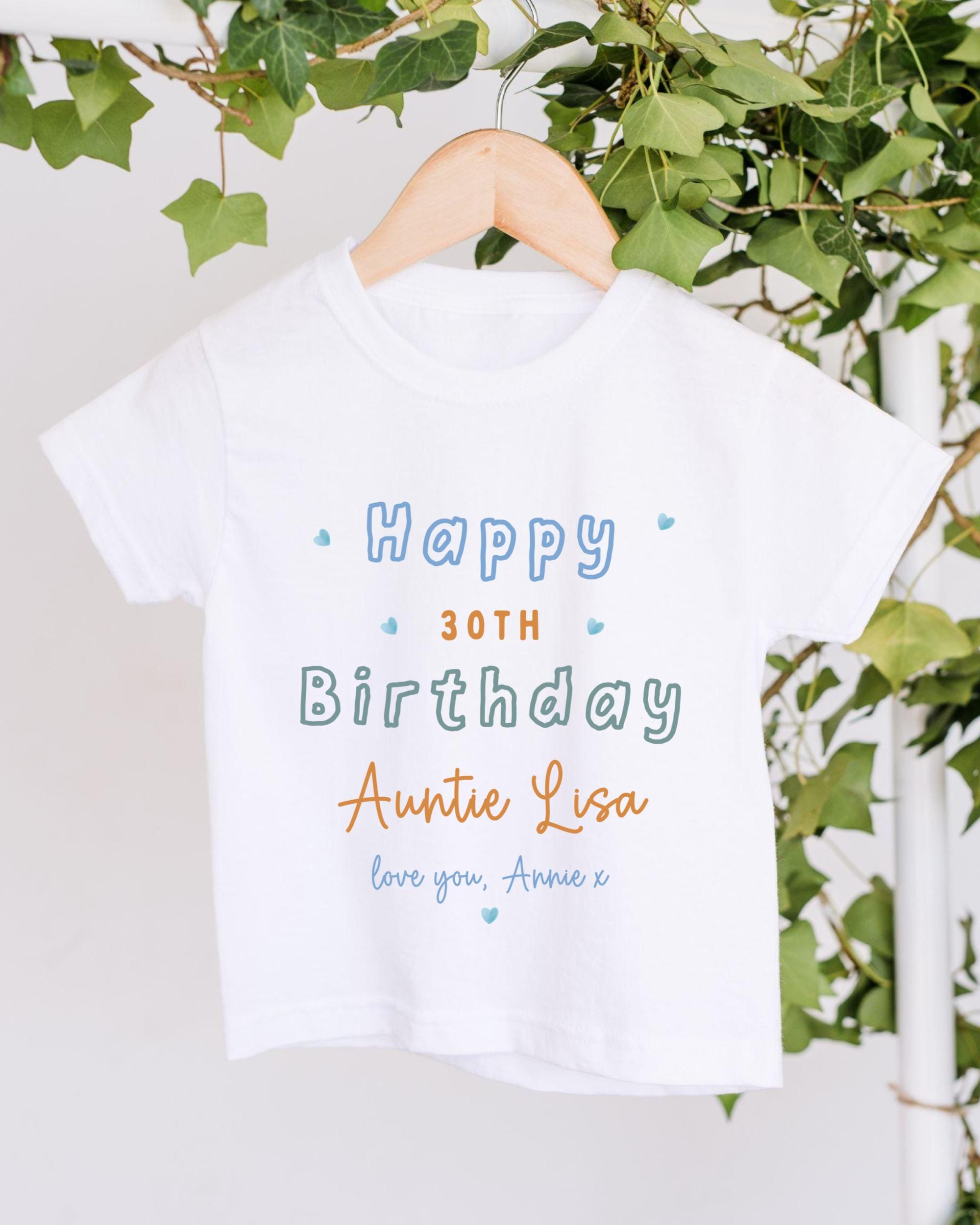 A white t-shirt with a birthday message printed on it, hanging on a wooden hanger.