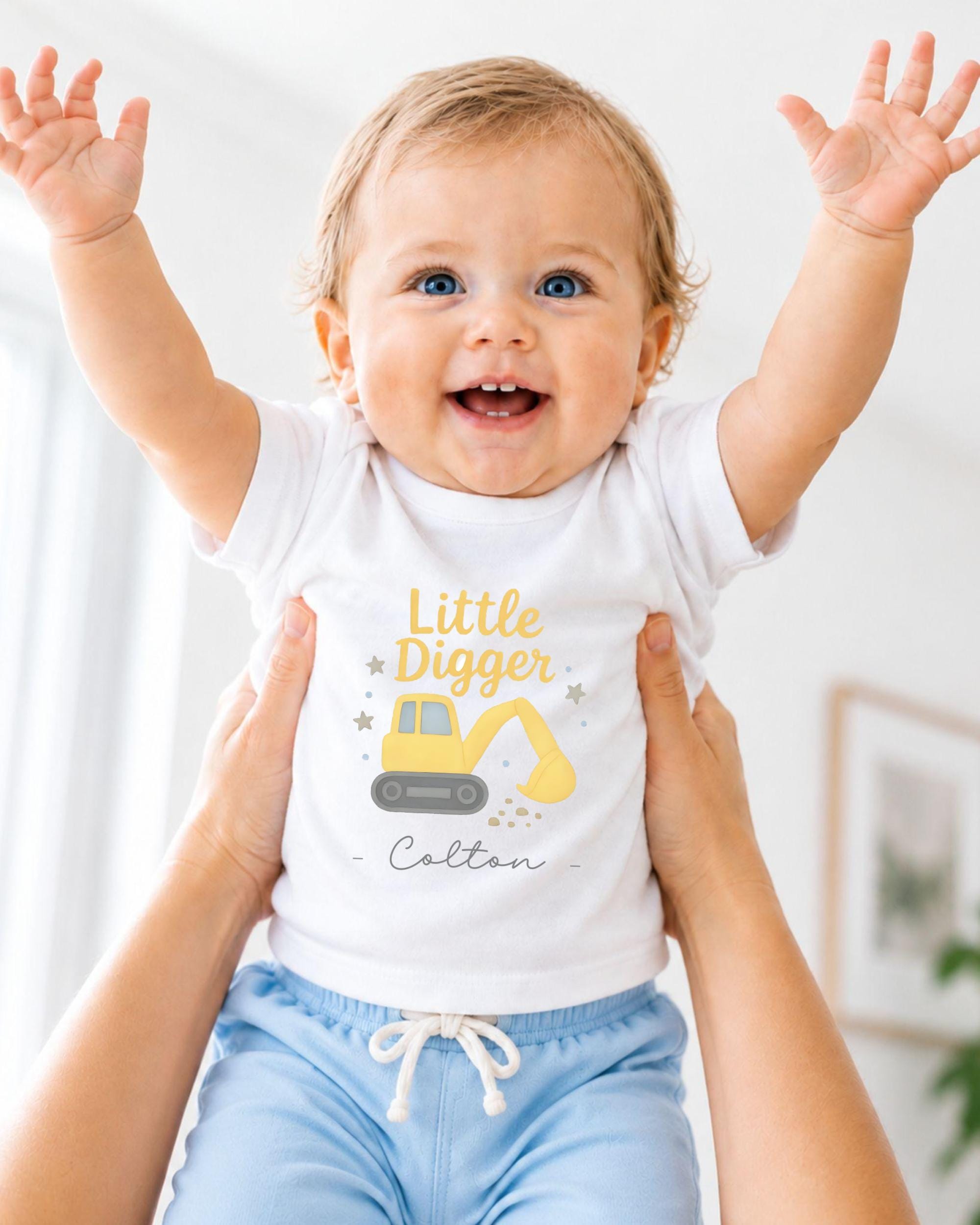 A smiling baby wearing a white t-shirt with a yellow construction vehicle graphic and the name "Colton" printed on it, being held up by someone's arms.