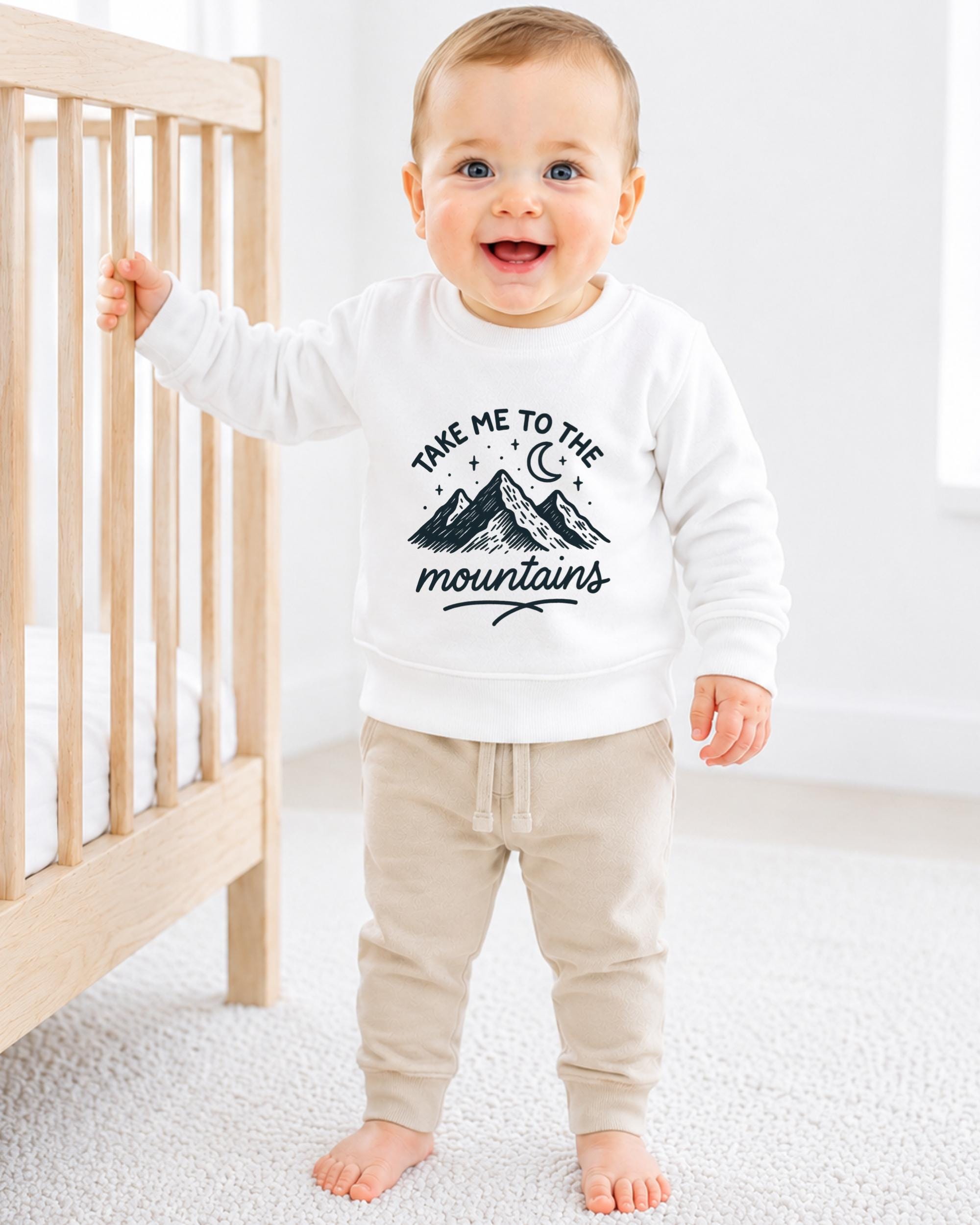 A smiling baby boy wearing a white sweatshirt with a mountain graphic and the text "Take me to the mountains" is standing next to a wooden crib.