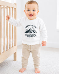 A smiling baby boy wearing a white sweatshirt with a mountain graphic and the text "Take me to the mountains" is standing next to a wooden crib.