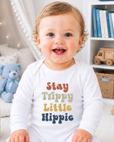 A smiling baby wearing a white onesie with the text "Stay Trippy Little Hippie" printed on it.