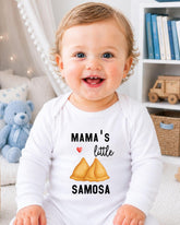 A smiling baby wearing a white onesie with a printed design of samosas and the text "Mama's little samosa" is sitting in a room with a teddy bear and bookshelf in the background.