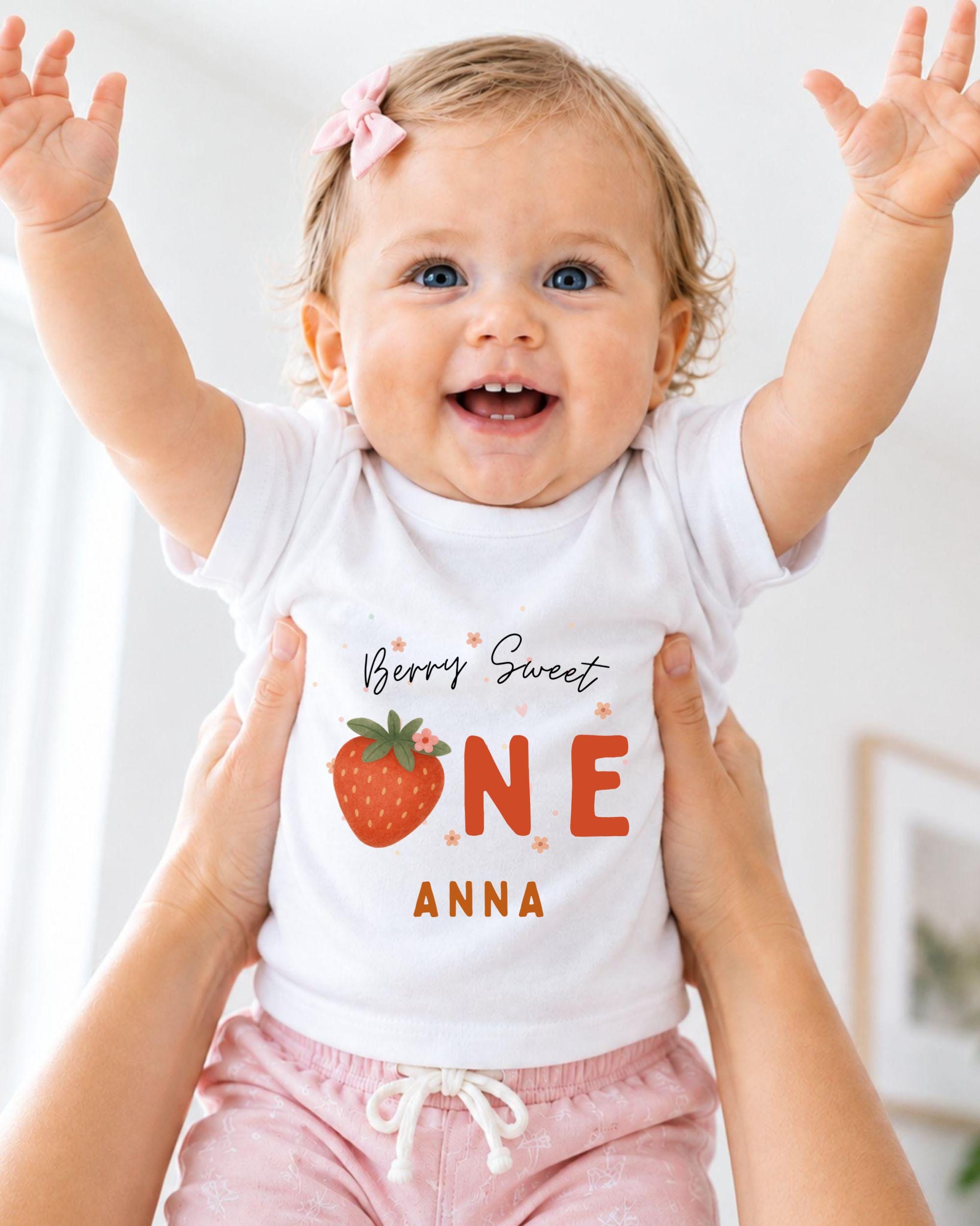 A smiling baby girl wearing a white t-shirt with a strawberry design and the text "Berry Sweet One Anna" is being held up by someone's arms.