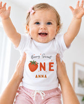 A smiling baby girl wearing a white t-shirt with a strawberry design and the text "Berry Sweet One Anna" is being held up by someone's arms.