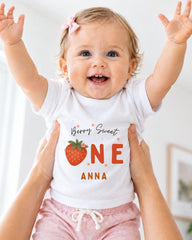 A smiling baby girl wearing a white t-shirt with a strawberry design and the text "Berry Sweet One Anna" is being held up by someone's arms.