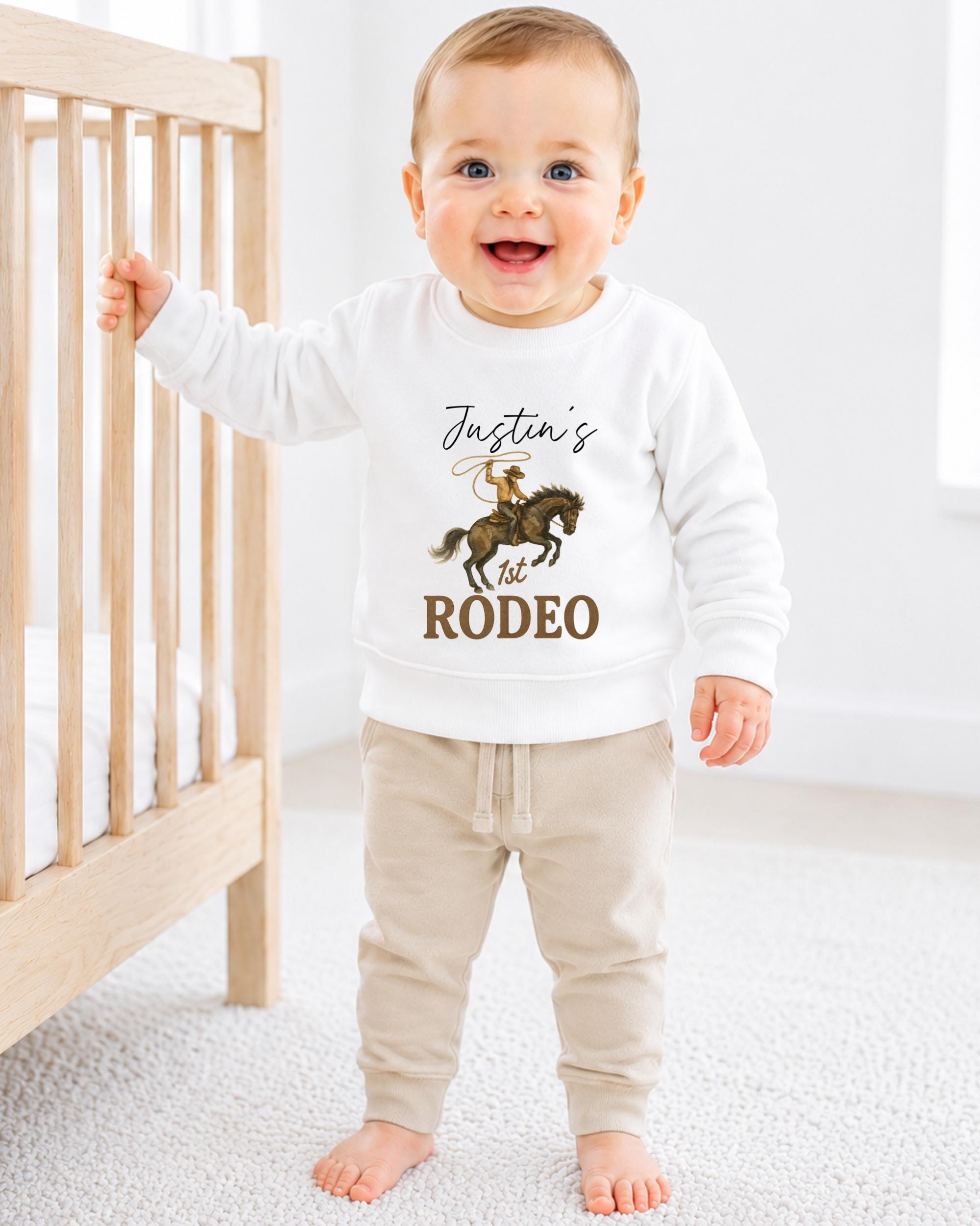 A smiling baby boy wearing a white sweatshirt with a cowboy graphic and the text " Justin's 1st Rodeo" is standing next to a wooden crib.
